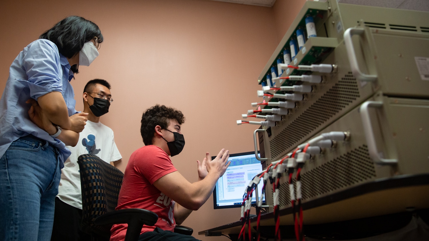 Mengmeng Zhu and Rui Wang stand Aidan Lim (seated in office chair) as they all look at results on a computer monitor. In the foreground a giant tan machine with batteries and red and white cables is visible.