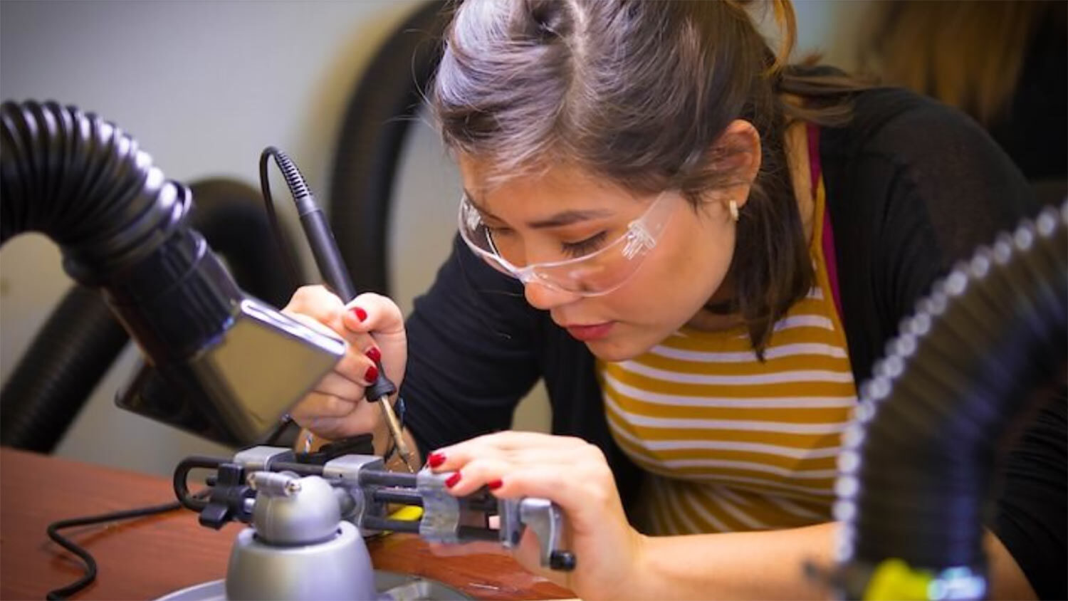 A student wearing safety goggles solders a piece of metal.